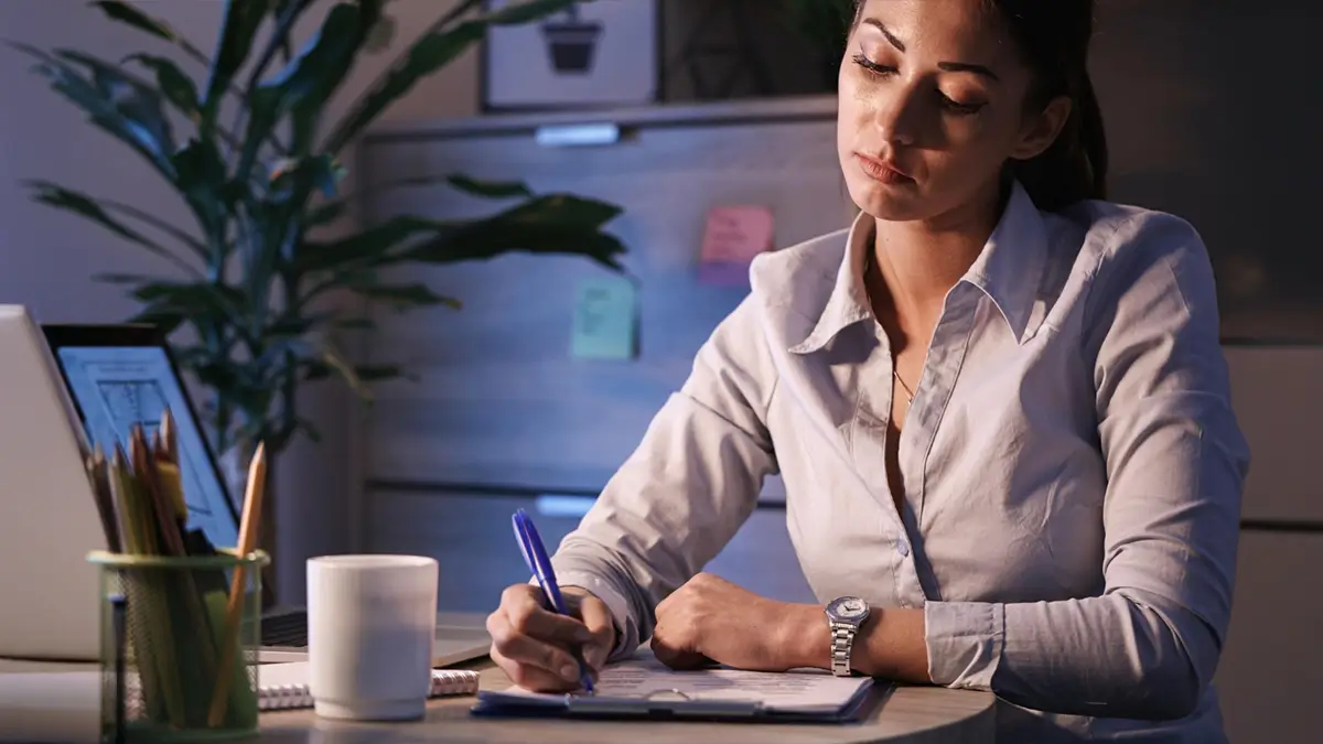 Uma mulher jovem sentada em uma mesa de escritório à noite, com foco nela escrevendo em uma prancheta com uma caneta. Há um notebook e material de escritório desfocados em primeiro plano e um armário com post-its e uma planta ao fundo. Representa a Governança para instituições sem fins lucrativos.